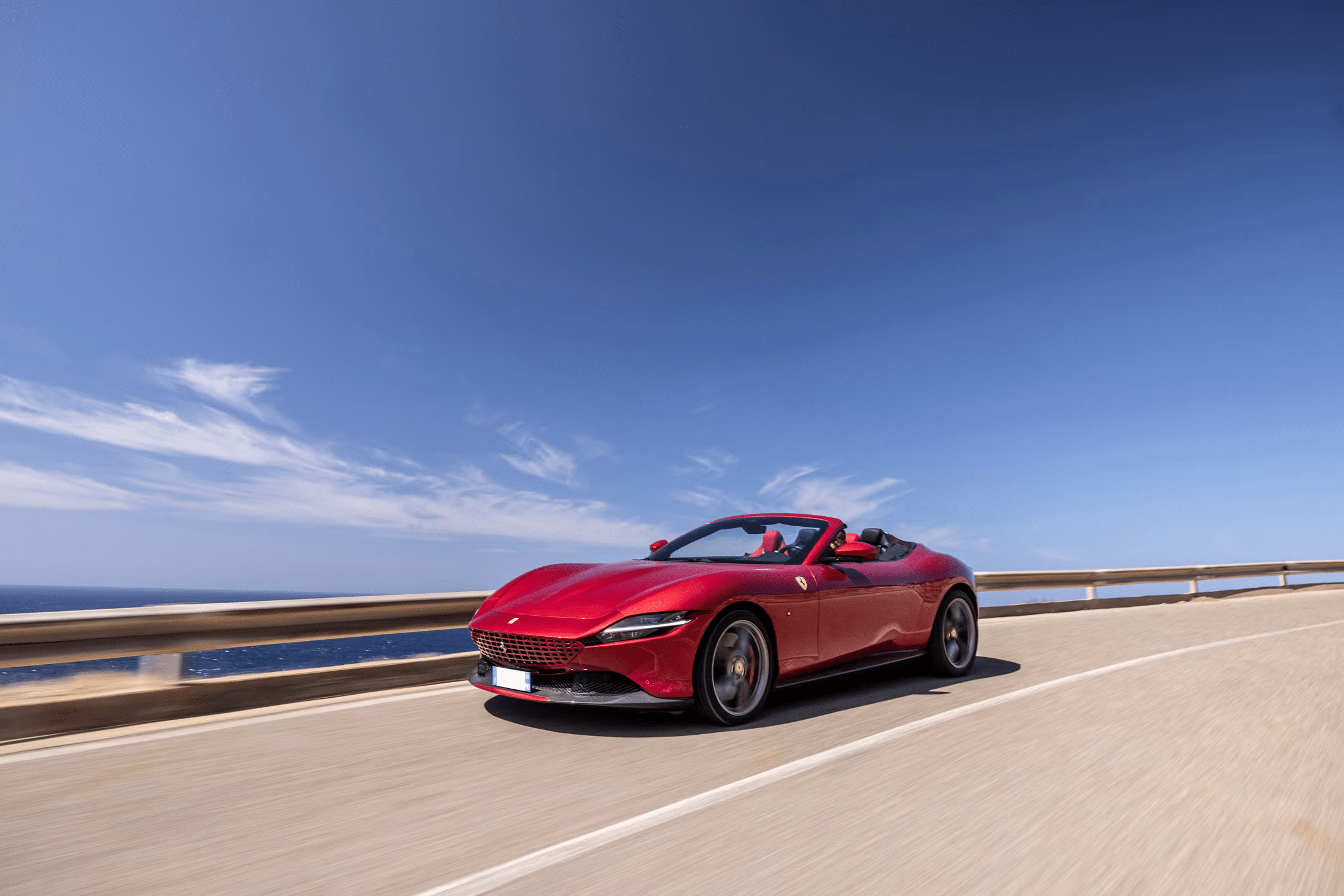 Red Ferrari Roma Spider front three-quarter view driving on a coastal highway with the soft top retracted and ocean in the background
