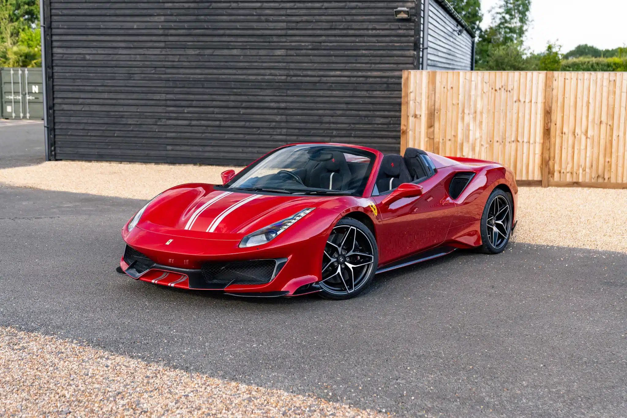 Rosso Berlinetta Ferrari 488 Pista Spider front three-quarter view parked on gravel with black timber building in background